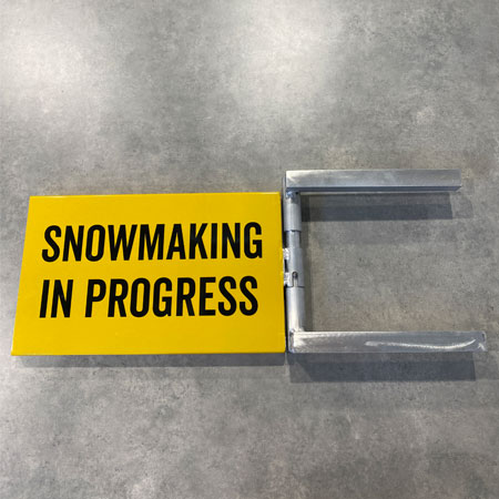Aluminum trail sign extender in the extended size with a locking mechanism on the left to secure in place for ski resorts. This image is in green and on white text is written Snowmaking In Progress